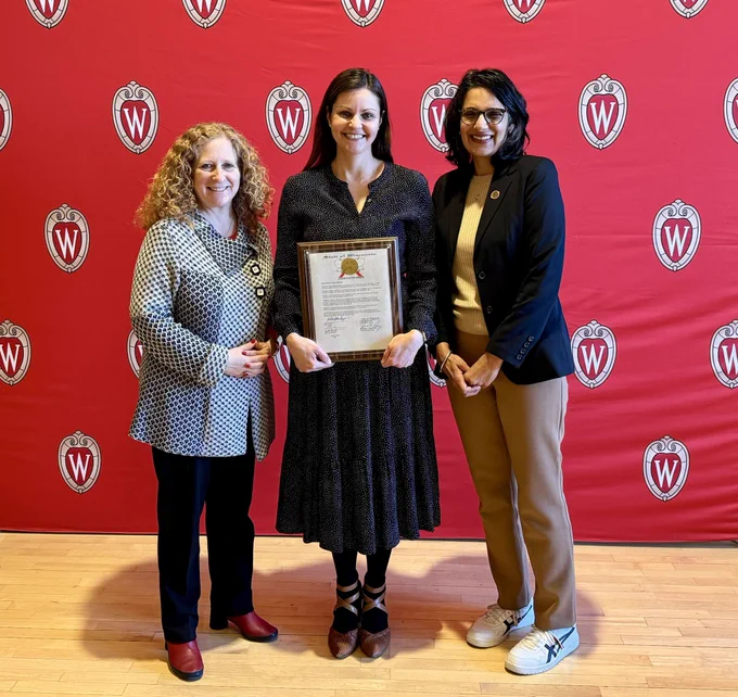 Chancellor Mnookin and Rep. Renuka Mayadev on either side of DTA awardee Claus Elholm Andersen's wife smiling in front of a red Wisconsin crest backdrop.