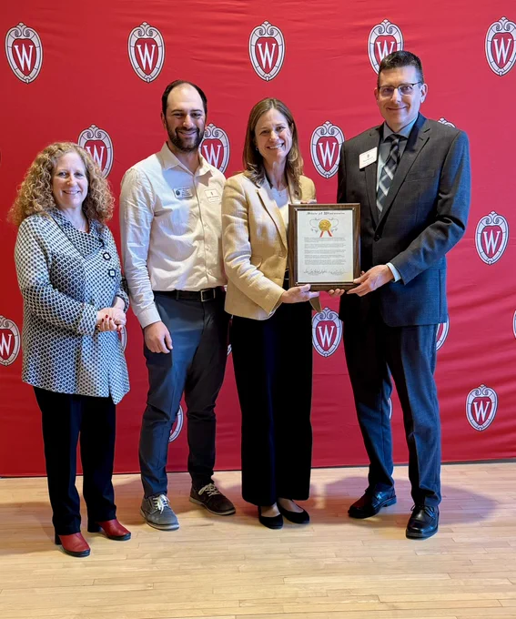 Chancellor Mnookin, Robby Abrahamian (chief of staff to Sen. Jodi Habush-Sinykin), Rep. Deb Andraca, and DTA Awardee Paul Roback smiling in front of a red Wisconsin crest backdrop.