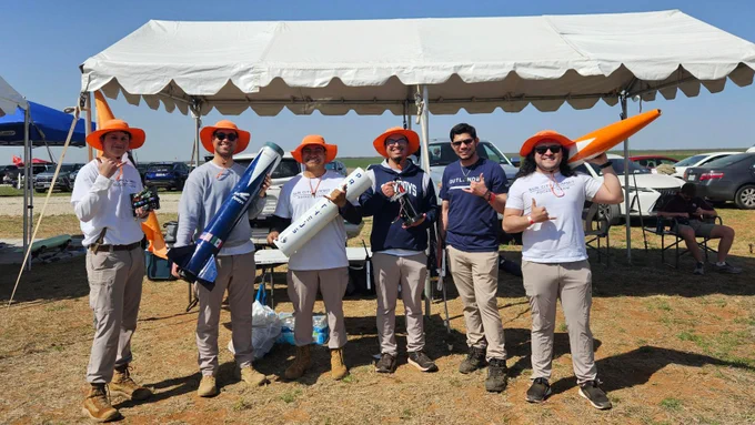 Six members of the University of Texas at El Paso's Sun City Summit Rocket Team posing with a rocket under a white tent, all wearing orange hats and UTEP shirts.