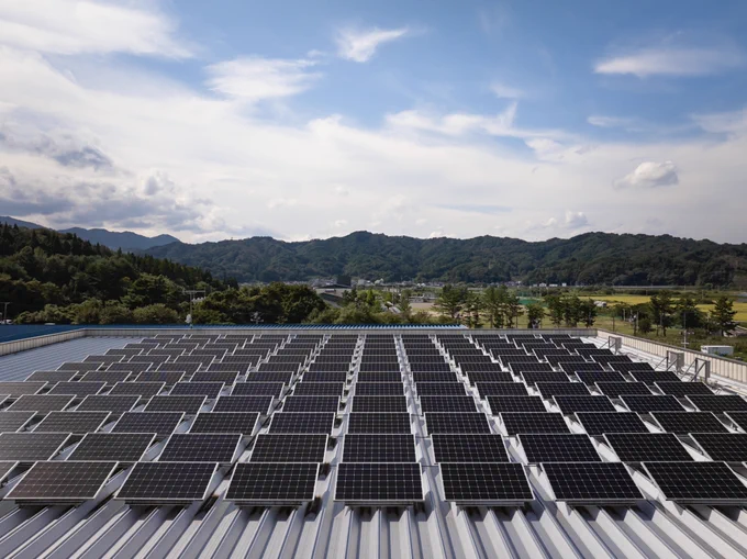 Multiple rows of solar panels are mounted on a rooftop, facing a partly cloudy sky. In the background, there is a forest of green trees on a mountain range and a valley of trees and buildings.