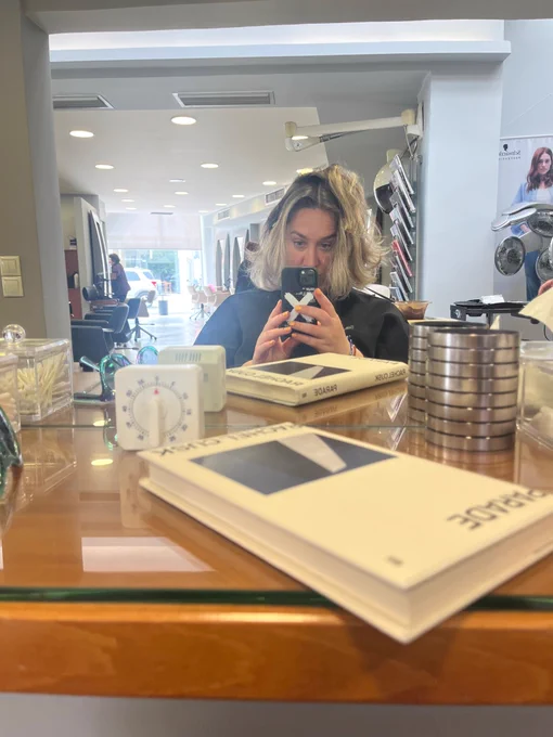 a blonde woman is taking a seated selfie in front of a hairdresser’s mirror. in front of her, Rachel Cusk’s book with the title Parade is visible.behind her one can see the hair salon (& chairs, spotlights, mirrors)