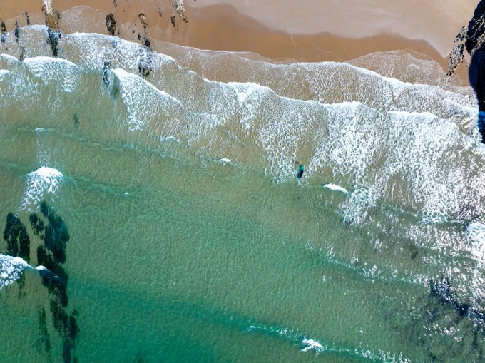 Aerial view of a beach with clear turquoise water gently lapping against the sandy shore. The waves create white foam as they break, and dark patches of seaweed or rocks are visible beneath the water's surface. The shoreline curves slightly, and there is a person in the water near the centre of the image.