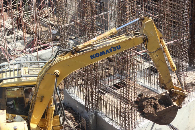 Excavator with a yellow arm is digging at a construction site. In the background, steel rebar frameworks are visible, indicating ongoing structural work.