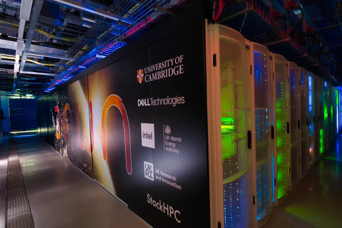 A panoramic view of the Dawn supercomputer servers. The wall prominently displays logos, including the University of Cambridge, Dell Technologies, Intel, UK Atomic Energy Authority, UK Research and Innovation, and SlackHPC.