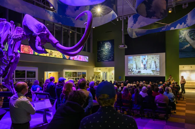 a mammoth skeleton is dramatically lit with purple at the back of a gallery filled with seated people watching two people present photos on a large screen