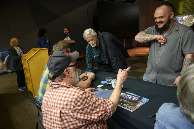 a person seated at a table is talking with a standing person about the sleeve of tattoos on their arm
