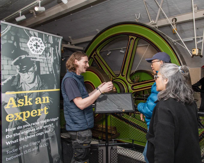 A member of the Technical team is talking to a couple in front of one of the flywheels and a poster in the Engine Rooms. 