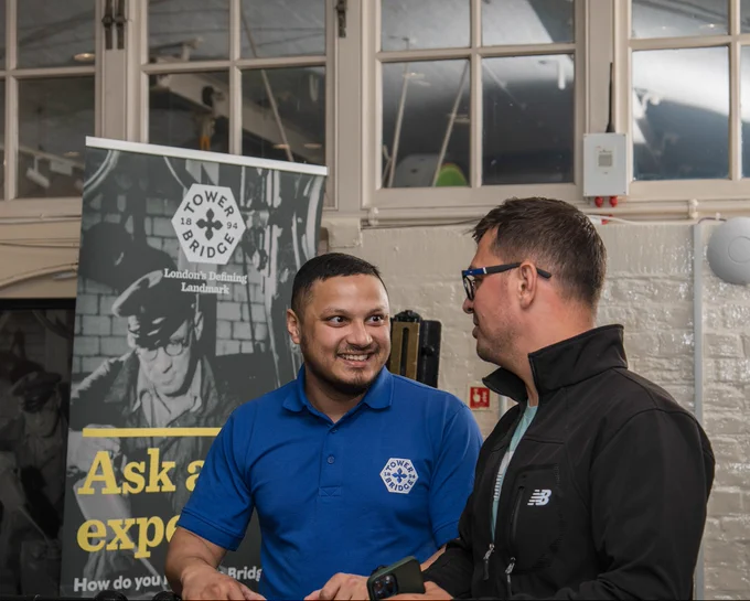 A member of the Technical team is talking to a person with glasses in front of a poster in the Engine Rooms. 