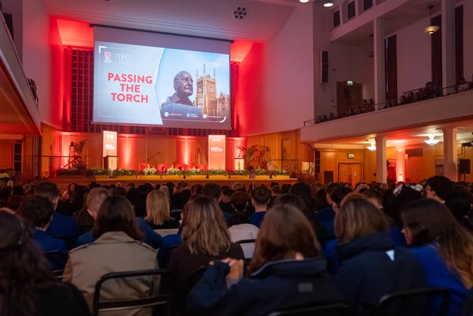 Image shows the audience looking towards the stage from the back of the room. Above the stage there is a screen with 'PASSING OF THE TORCH' artwork, which contains a photograph of the bust of Senator Mitchell which faces the Whitla Hall.