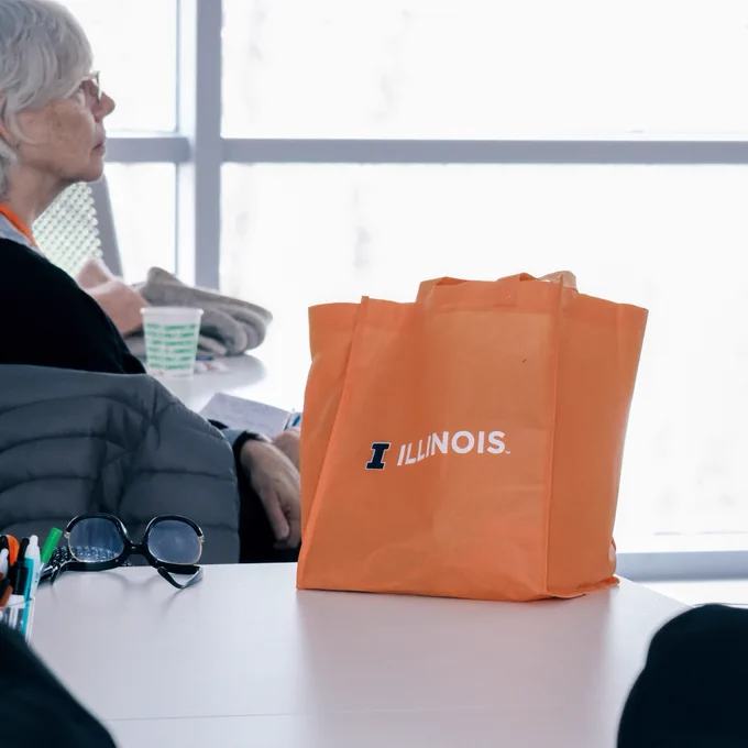 An orange bag featuring the Illinois block "I" logo followed by "Illinois" written in white across the center of the bag sits atop a table inside the Sunrise Studio (Room 1040) at the Siebel Center for Design on the University of Illinois Urbana-Champaign campus. 