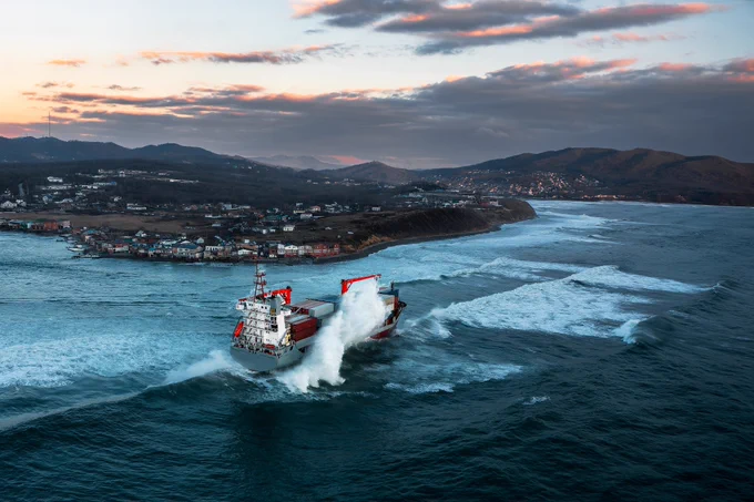 A cargo ship moves through ocean waves near the shore, with water crashing against the front of the vessel. In the background, a coastal town sits beneath a sunset sky, with hills and scattered houses visible.