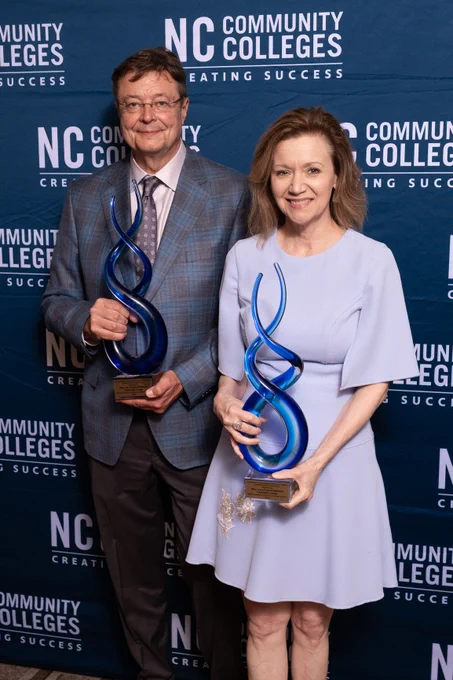 Two individuals holding awards at an NC Community Colleges event, standing in front of a banner with the organization's logo.