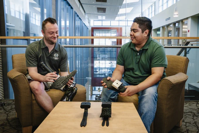 Two students sitting on sofa chairs, holding prosthetic feet, smiling. Placed on a table in front of them are two models of the internal structure of the prosthetic feet.