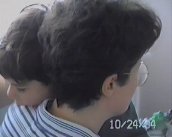 Holly Márie Parnell, still from Cabbage (2023). Courtesy of the artist. [Image description: A camcorder still showing a close-up of brown-haired child draping their head over a parent's shoulder. The parent, who also has short brown hair, faces away from the camera and wears glasses and a striped collared shirt. The date October 24 1994 appears in the bottom right corner. ]