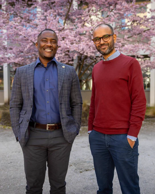 Kwadwo Oti-Sarpong and Terry de Sousa smiling and standing in front of blossoming cherry trees. Kwadwo is on the left, wearing a blazer and buttoned shirt. Terry, on the right, is in a red sweater and glasses.