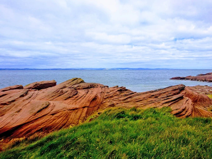 Permian desert sandstone on the north Arran coast. In the foreground is grass and in the background is the sea. It is a cloudy day. 