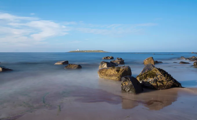 Pladda island from Kildonan at the south of the island. You can see a small amount of mist across the sand at the forefront of the image, and some large rocks are dotted around. It is a sunny day, with some small clouds off to the left. 