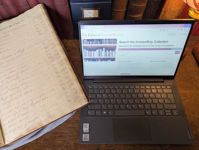 A desk with two versions of a library catalogue, an open, hand-written book and a laptop computer showing a search page.