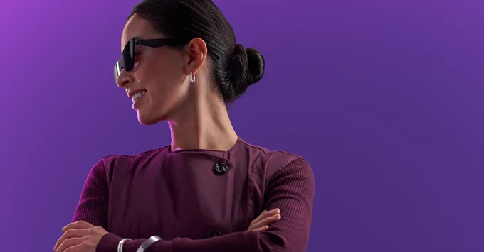 A woman with sunglasses, a purple shirt, and dark hair, smiles. The background is also purple.