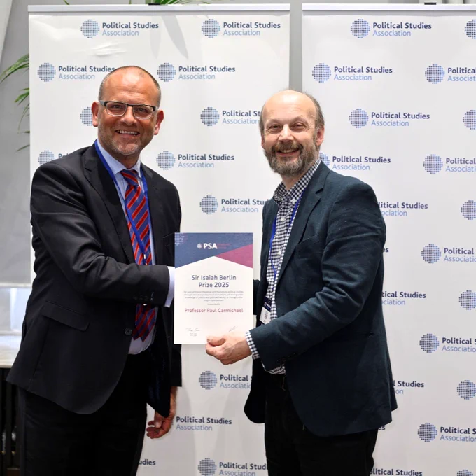 Two men in suits smile and shake hands while one hands an award certificate to the other. They stand in front of a backdrop with Political Studies Association logos.