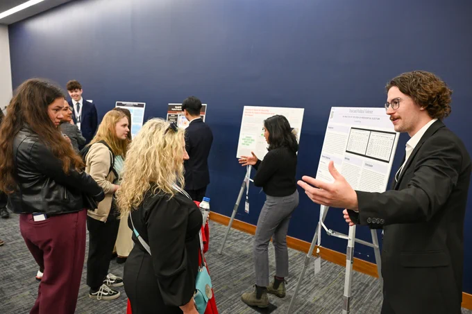 Individual presenting a poster to a group of attentive listeners at a professional conference. Several other people are engaged in discussions around adjacent posters.