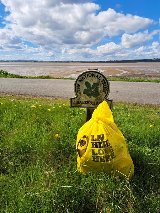 Full bag of litter lifted at Ballyreagh in front of the National Trust sign which is placed on the shore of Strangford Lough. The tide is out. Sky is blue with a scattering of white fluffy clouds 