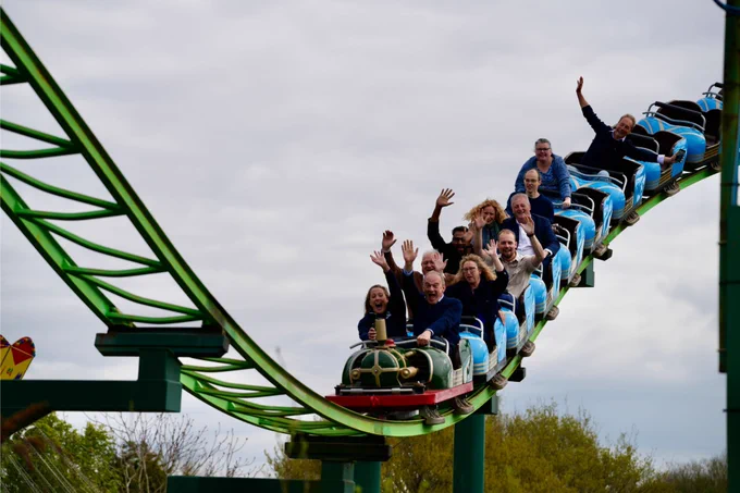 Ed Davey, Ian Roome MP, and a group of supporters ride a rollercoaster, hands in the air.