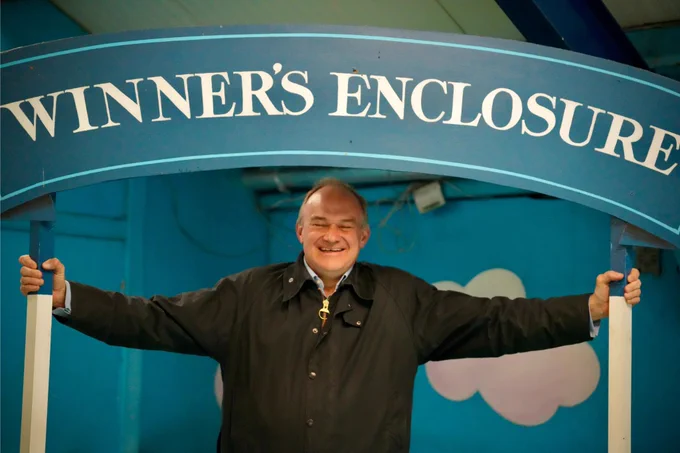 Ed Davey stands grinning under a blue archway labelled “WINNER’S ENCLOSURE,” holding the frame with both hands.