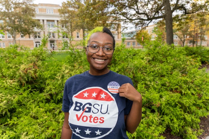 A person wearing a "BGSU Votes" t-shirt and holding an "I Voted" sticker, smiling in front of green shrubs with a building in the background.