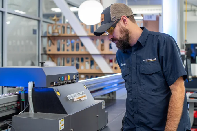 A man in a dark blue short sleeve button up stands in front of a gray printer machine. He wears glasses and a navy blue hat while looking down at the machine. In the background is a wooden wall of shelves.