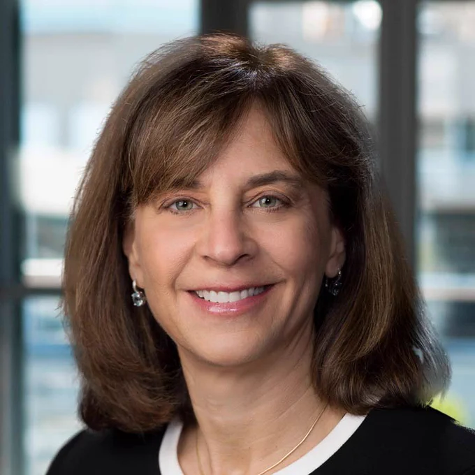 Portrait of a smiling person with shoulder-length brown hair, wearing a black blazer and a white top, set against an office background. 