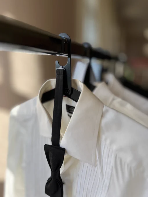 A crisp white tuxedo shirt hangs neatly on a hanger. The bow tie is perfectly placed in the hanger’s notch, creating a balanced and refined presentation. The shirt is sharply pressed, with clean lines that emphasize care and intention. A soft, neutral background allows the shirt and hanger to take center stage, with gentle lighting highlighting the fabric’s texture and the hanger’s minimalist design.