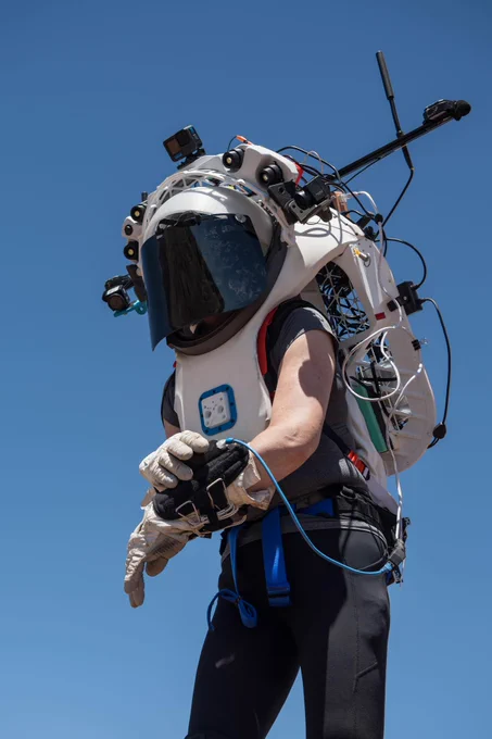 Person outdoors under a blue sky, wearing a partial mockup of an astronaut EVA suit, adjusting a device attached to the cuff of her wrist. The suit has a dark, tinted visor. Wires connect many points on the suit's periphery, and devices on various mounts extend from its skeletal structure. Photo credit: NASA/Josh Valcarcel