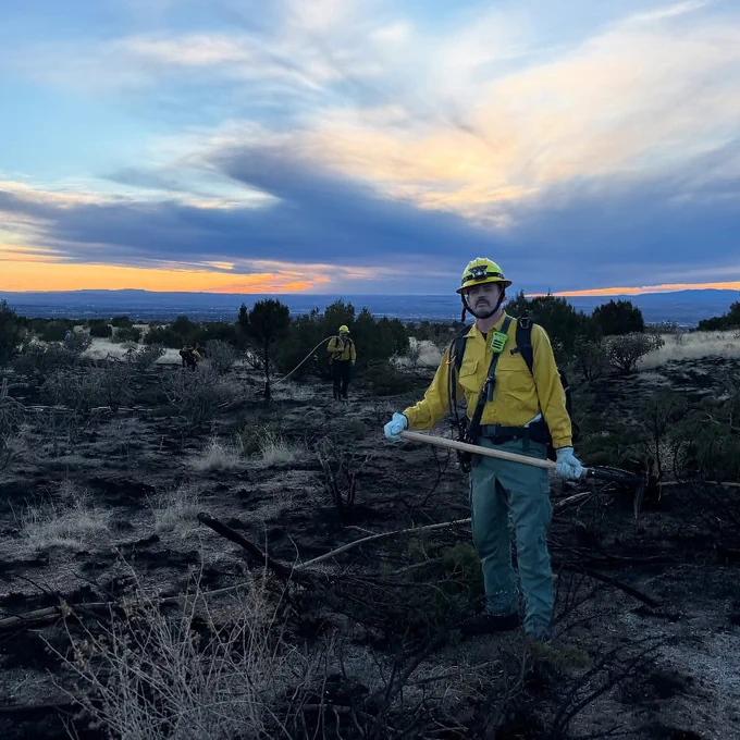 A firefighter, wearing yellow safety gear and holding a digging tool, poses in a recently burnt area of brush. The mesa and city are visible in the background behind him.