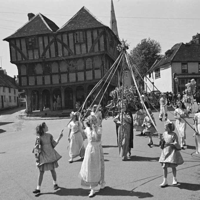 A black and white photograph of young children dancing around a small wooden maypole in front of a timber-framed building.