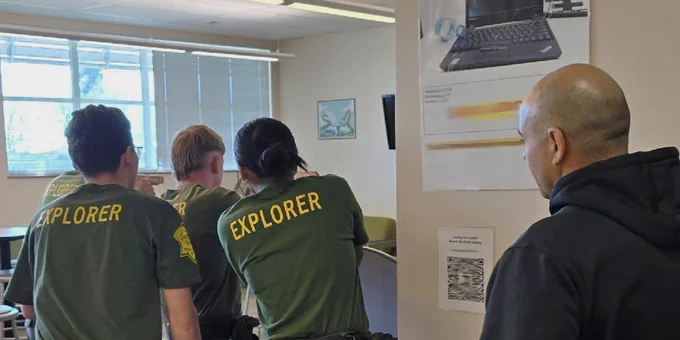 Three individuals in 'Explorer' uniforms round a corner to clear a room, observed by another person in a dark jacket.