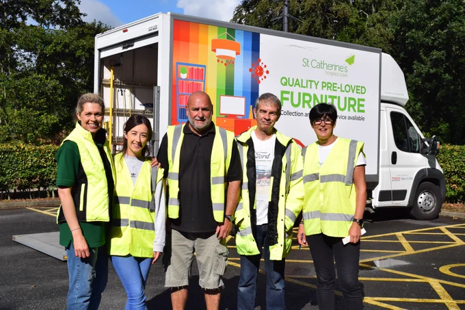 Four volunteers wearing hi vis jackets stood in front of a St Catherine's branded lorry which has 'St Catherines hospice care quality pre-loved furniture' written on the side