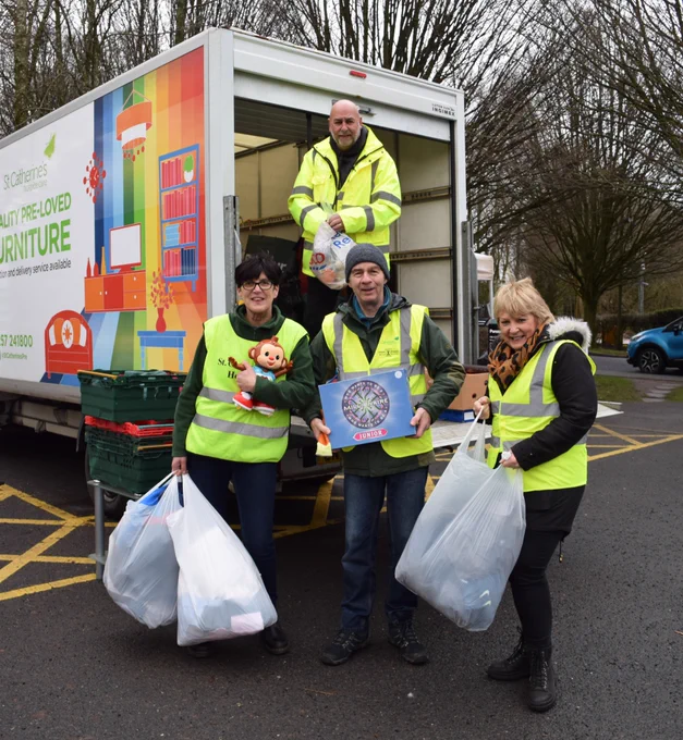 Four volunteers wearing hi vis jackets stood in front of a St Catherine's branded lorry holding pre-loved goods which have been donated