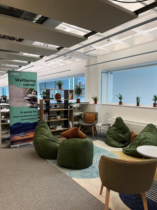 Photo of the Wellbeing Corner on level 03. Beanbags and comfortable chairs, cushions, a rug and a book case with books, ornaments and artificial plants. Pull up banner that reads 'Wellbeing corner. A space for silent meditation and contemplation.'