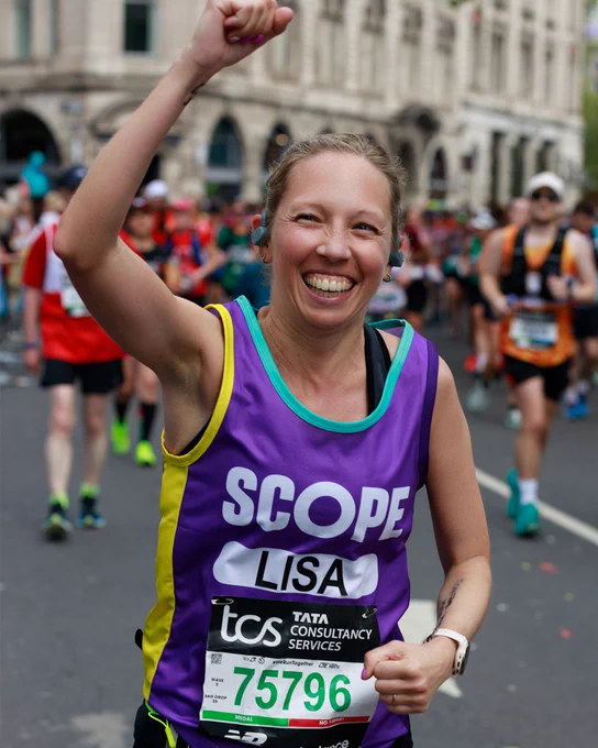 A photo of Lisa smiling widely. She is raising her fist in the air in celebration while she runs past the camera. She wears a bright purple Scope running vest.