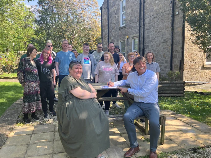a group of people is around a wooden table in a garden. sitting at the table is NIN member Lynnette with RAMH CEO Peter Jung. The sun is shining and people are smiling. Peter is signing the Charter certificate. 