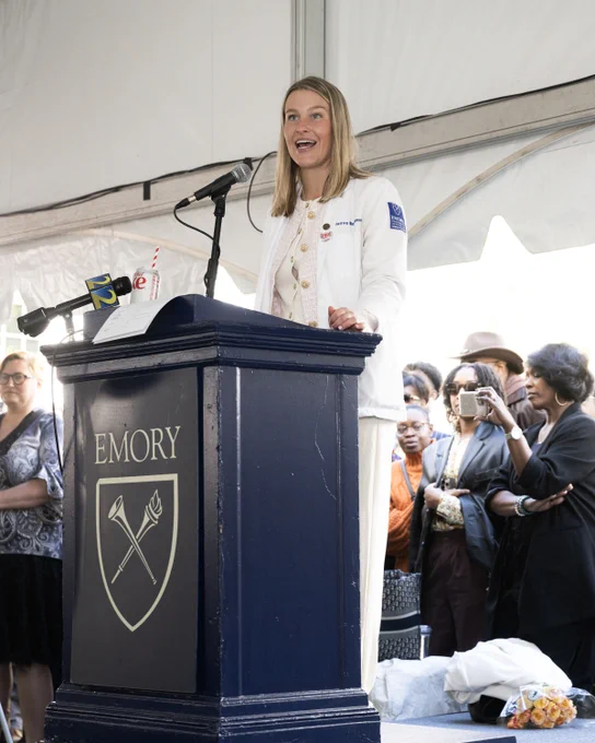Person speaking at a podium with the Emory University logo, surrounded by an audience under a white tent.
