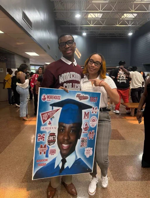 A student wearing a Morehouse College sweatshirt is standing in a school cafeteria holding a large sign printed with a graduation photo of himself surrounded by Morehouse logos. He is standing in a cafeteria next to a loved one who is pointing at him. They are both smiling at the camera. 