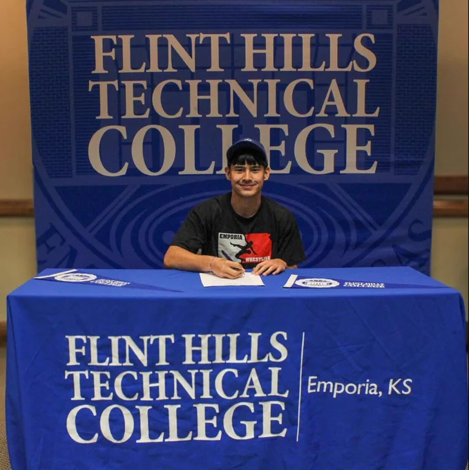 A student is sitting at the same Flint Hills Technical college table. He is wearing a blue hat and a black Emporia Wrestling shirt. He is smiling at the camera. 