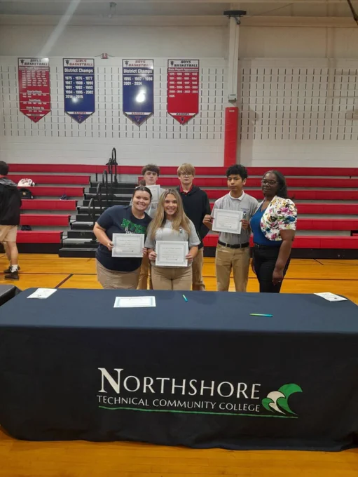 Five students are standing behind a table with a Northshore Technical Community College tablecloth in a school gym with an educator. The students are holding certificates in their hands and smiling at the camera. 
