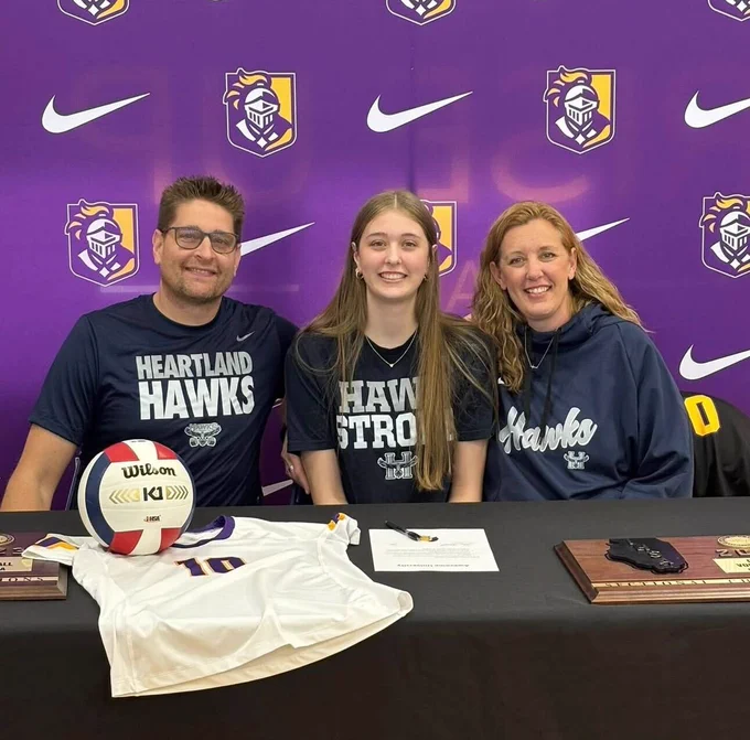 A student and their parents are sitting behind a table that has a volleyball and an athletic jersey on it. They are all wearing Heartland Hawks merch and are smiling at the camera. 