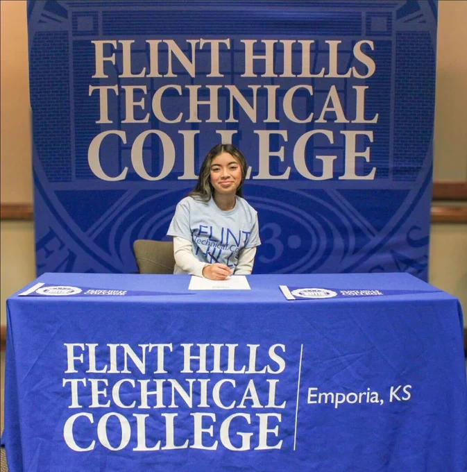 A student is sitting behind a table with a Flint Hills Technical College tablecloth. She is smiling a the camera and wearing a light blue shirt with the college’s logo on it. 