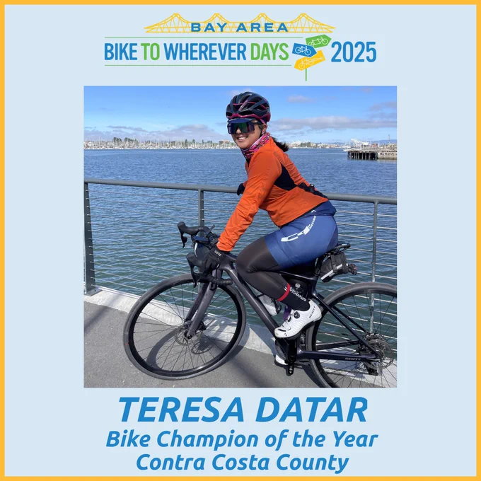 Photo of a woman on a black road bike wearing a black bike helmet, aerodynamic sunglasses, blue bike shorts, black bike leggings, and white bike shoes with the Bay in the background.