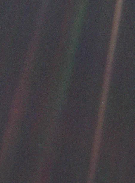 A faint image showing Earth as a tiny pale blue dot, less than a pixel in size, appearing in a scattered ray of sunlight against the dark backdrop of space. Captured by NASA’s Voyager 1 spacecraft on February 14, 1990, from a distance of about 3.7 billion miles, the image emphasizes Earth's smallness amid vast space.