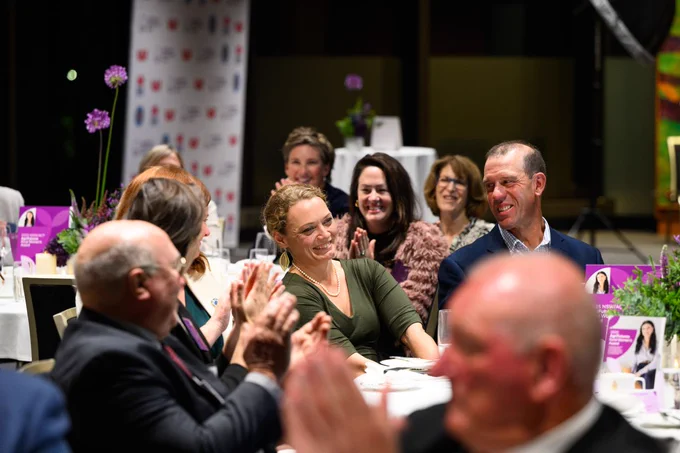People seated at a dinner event, enjoying a conversation and clapping as Carol Mudford (Winner is announced, with floral centerpieces on tables in a dimly lit room.